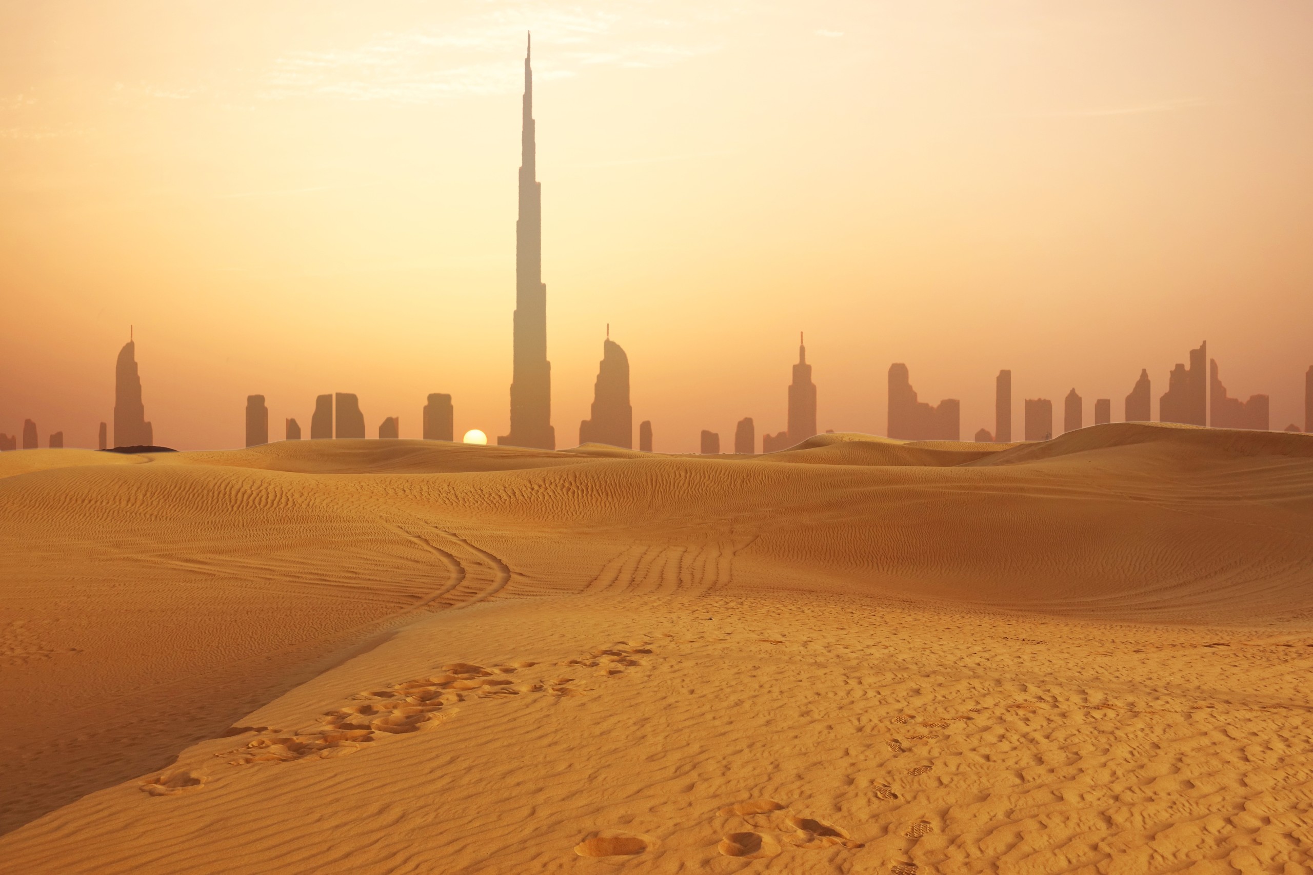 Desert sand dunes in the foreground with a city skyline and the Burj Khalifa tower in the background at sunset.