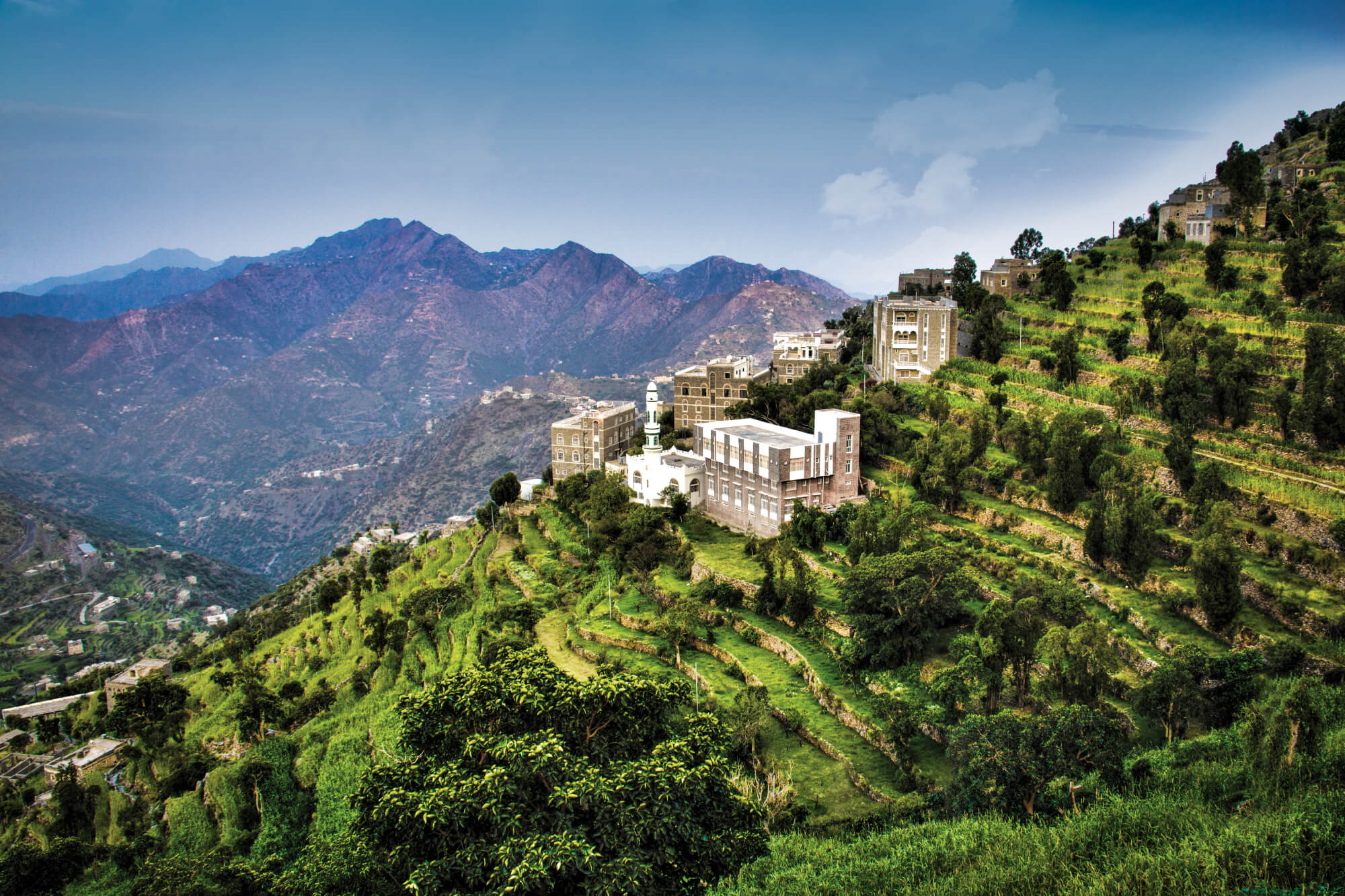 Terraced green hills with buildings scattered on slopes, mountains in the background, and a partly cloudy blue sky above.