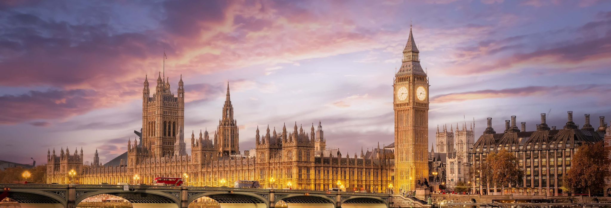 The Houses of Parliament and Big Ben in London are seen at sunset, with Westminster Bridge and a red double-decker bus in the foreground.