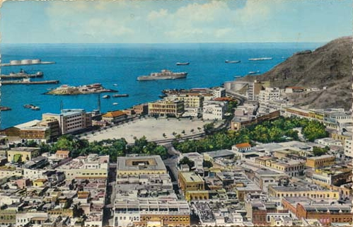 A coastal cityscape with a central open square, surrounding buildings, a waterfront, several ships docked at the port, and a hillside to the right.